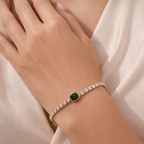 A close-view of a gold-plated tennis chain bracelet for women featuring a row of clear crystals and a central emerald-green stone
