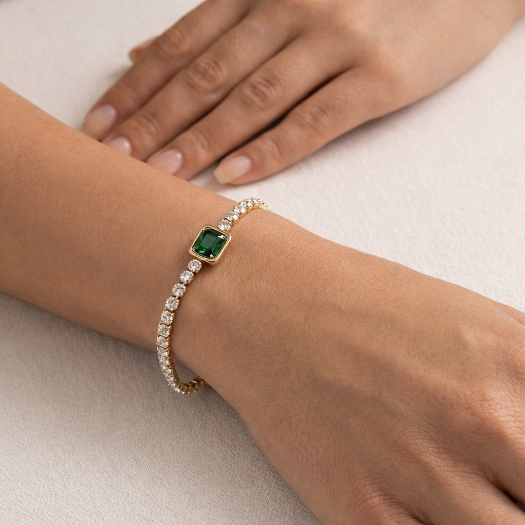 A gold-plated tennis chain bracelet for women featuring a row of clear crystals and a central emerald-green stone, shown on a model hand