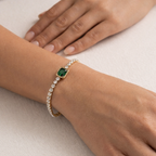 A gold-plated tennis chain bracelet for women featuring a row of clear crystals and a central emerald-green stone, shown on a model hand