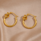 A pair of gold-plated hoop earrings for women featuring a central love-knot design, shown on a soft champagne silk background.