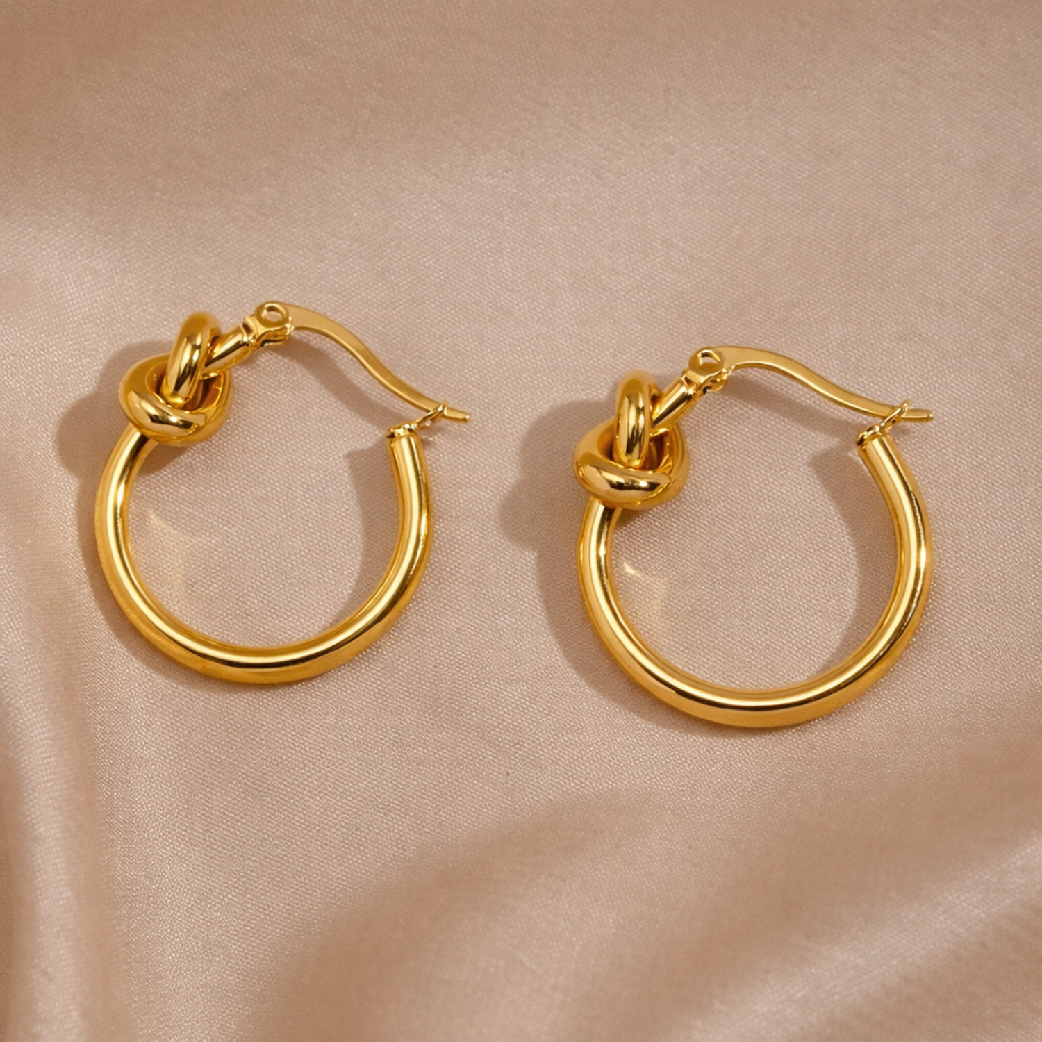 A pair of gold-plated hoop earrings for women featuring a central love-knot design, shown on a soft champagne silk background.