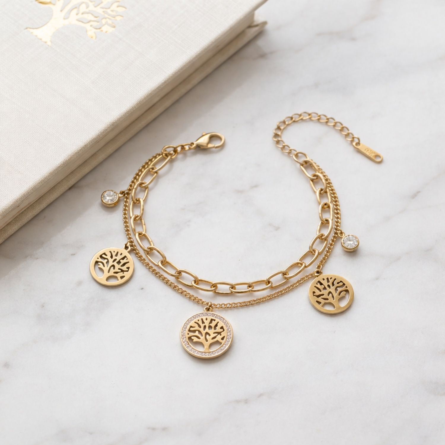A dual-layer gold-plated chain bracelet for women featuring Tree of Life charms and clear crystal accents, displayed on a marble surface next to a white book