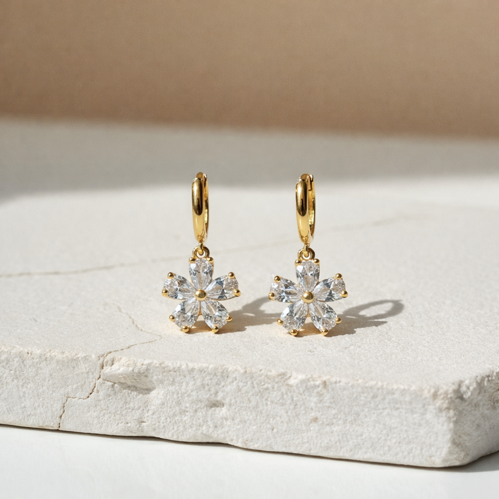 A pair of gold-plated hoop earrings for women featuring a sparkling five-petal crystal flower drop, shown on a white textured stone.