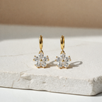 A pair of gold-plated hoop earrings for women featuring a sparkling five-petal crystal flower drop, shown on a white textured stone.