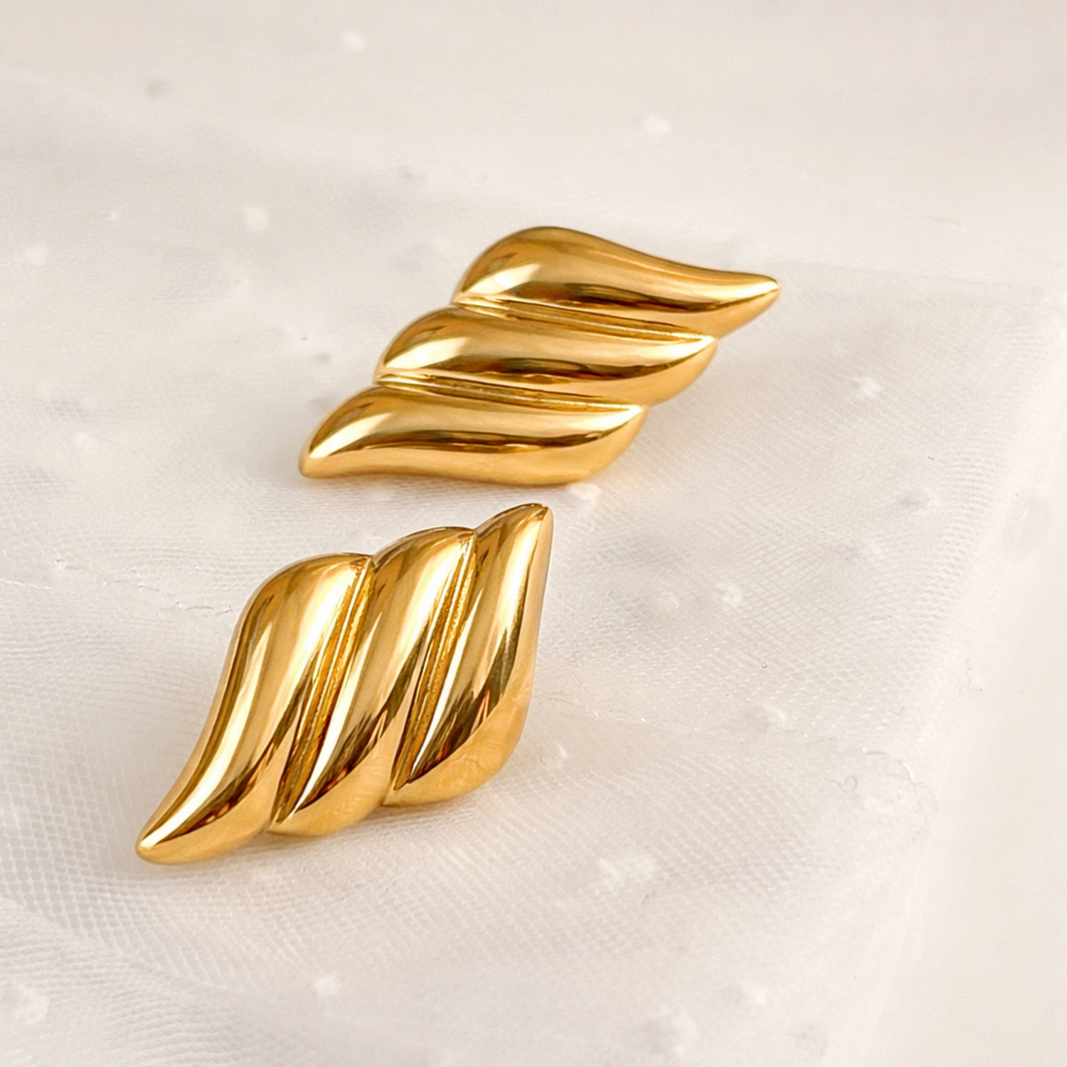 A pair of a gold-plated sculptural wing-shaped stud earring for women, shown on a textured surface