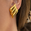 A close-up of a gold-plated sculptural wing-shaped stud earring for women, shown worn on a model's ear