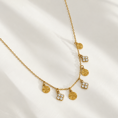 A gold chain necklace with five dangling charms, including two sunburst disc charms and three flower-shaped charms with clear rhinestones, displayed on a white fabric background with soft shadows.