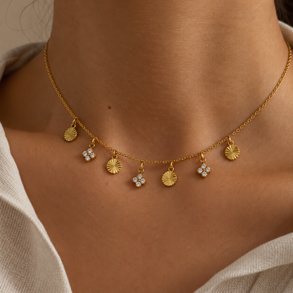 A close-up of a delicate gold chain necklace worn on a model's neck, featuring small dangling charms including sunburst and flower motifs, some embellished with clear stones.