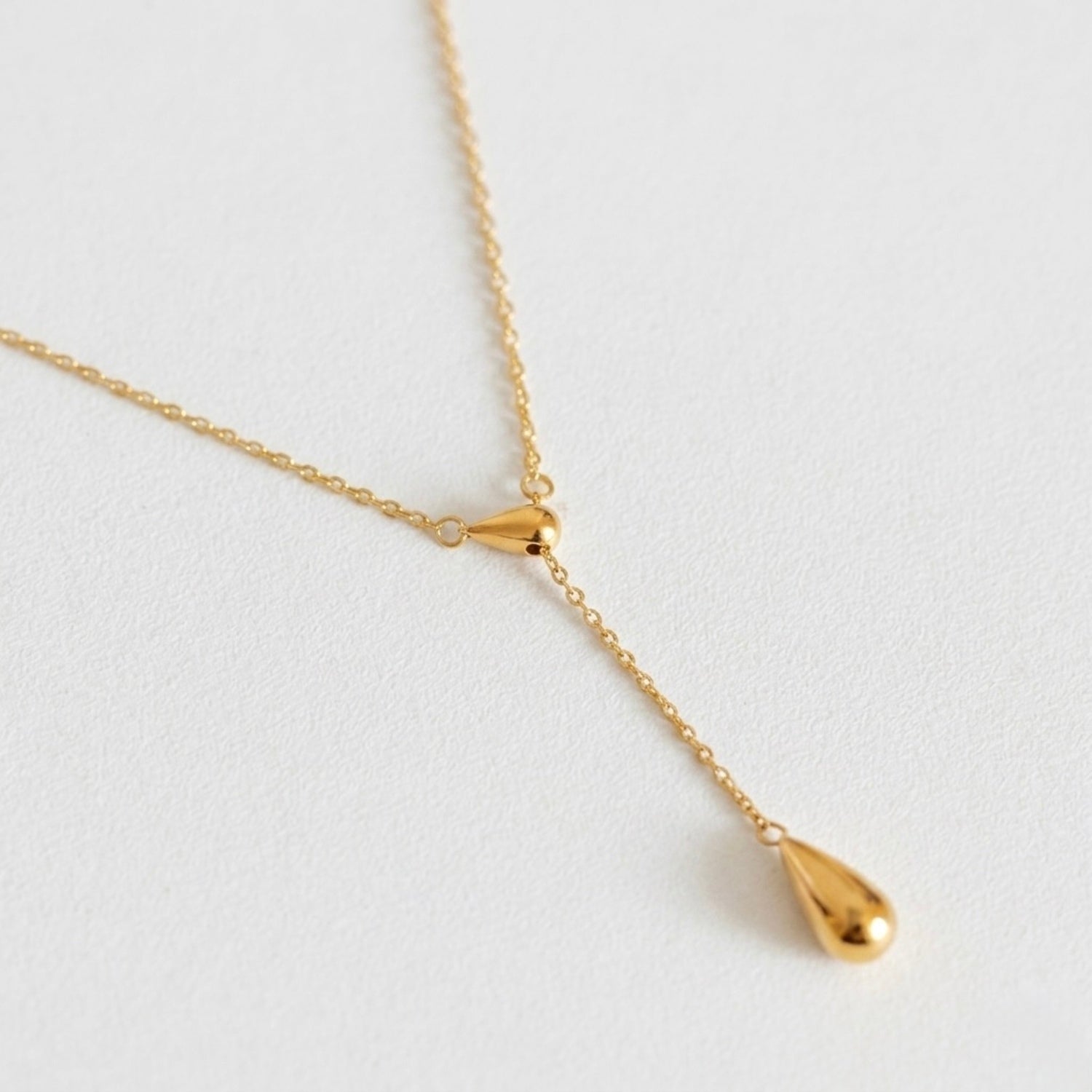 A gold-plated stainless steel chain for women featuring a Y-shaped lariat design with two polished teardrop-shaped pendants on a white textured background.