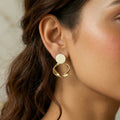 A gold-plated statement earrings for women featuring a flat circular stud and a sculptural twisted hoop pendant, shown on a model