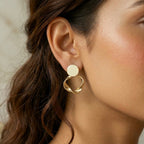 A gold-plated statement earrings for women featuring a flat circular stud and a sculptural twisted hoop pendant, shown on a model