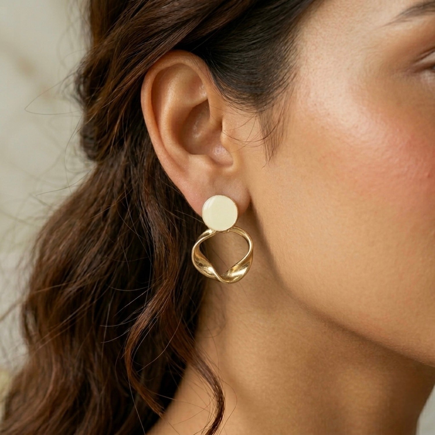 A gold-plated statement earrings for women featuring a flat circular stud and a sculptural twisted hoop pendant, shown on a model