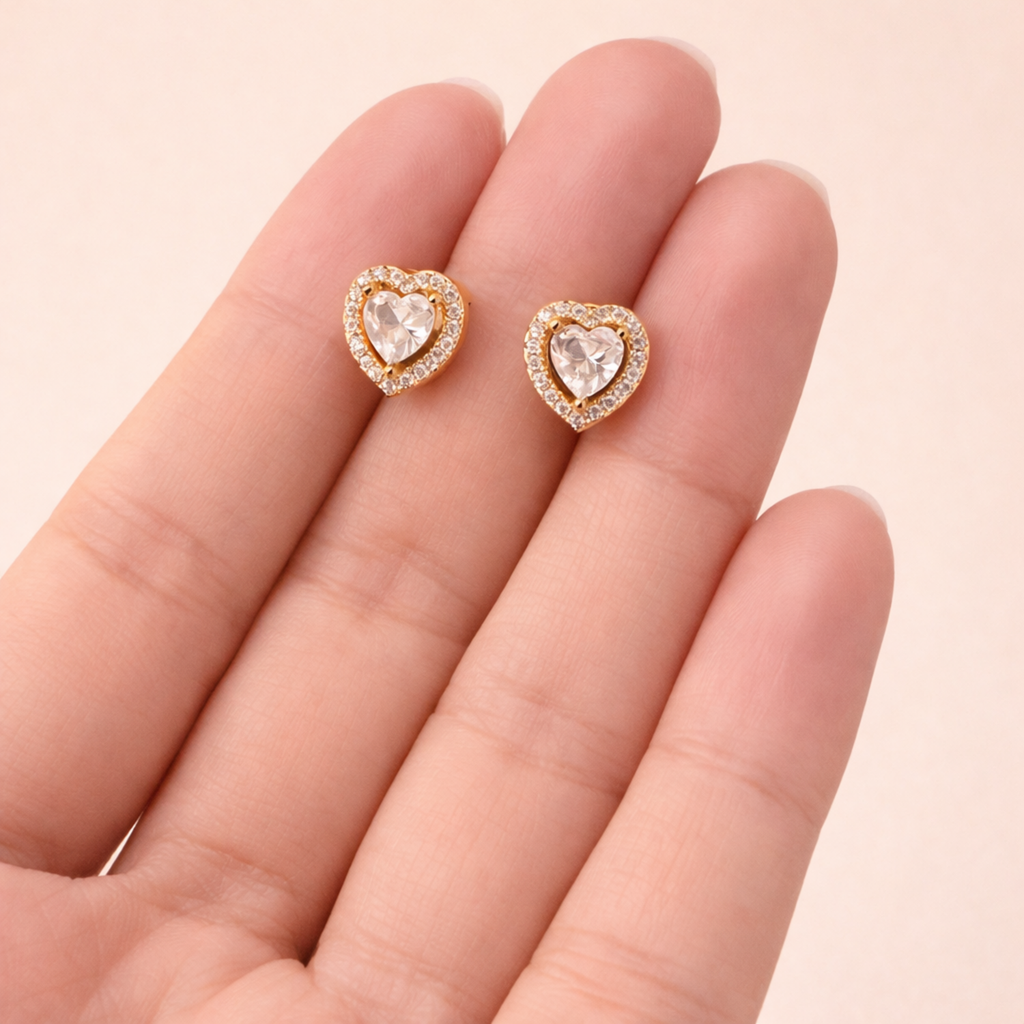 A pair of gold-plated heart-shaped stud earrings for women featuring a large central clear crystal and a pavé halo, shown held in a hand.