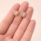 A pair of gold-plated heart-shaped stud earrings for women featuring a large central clear crystal and a pavé halo, shown held in a hand.