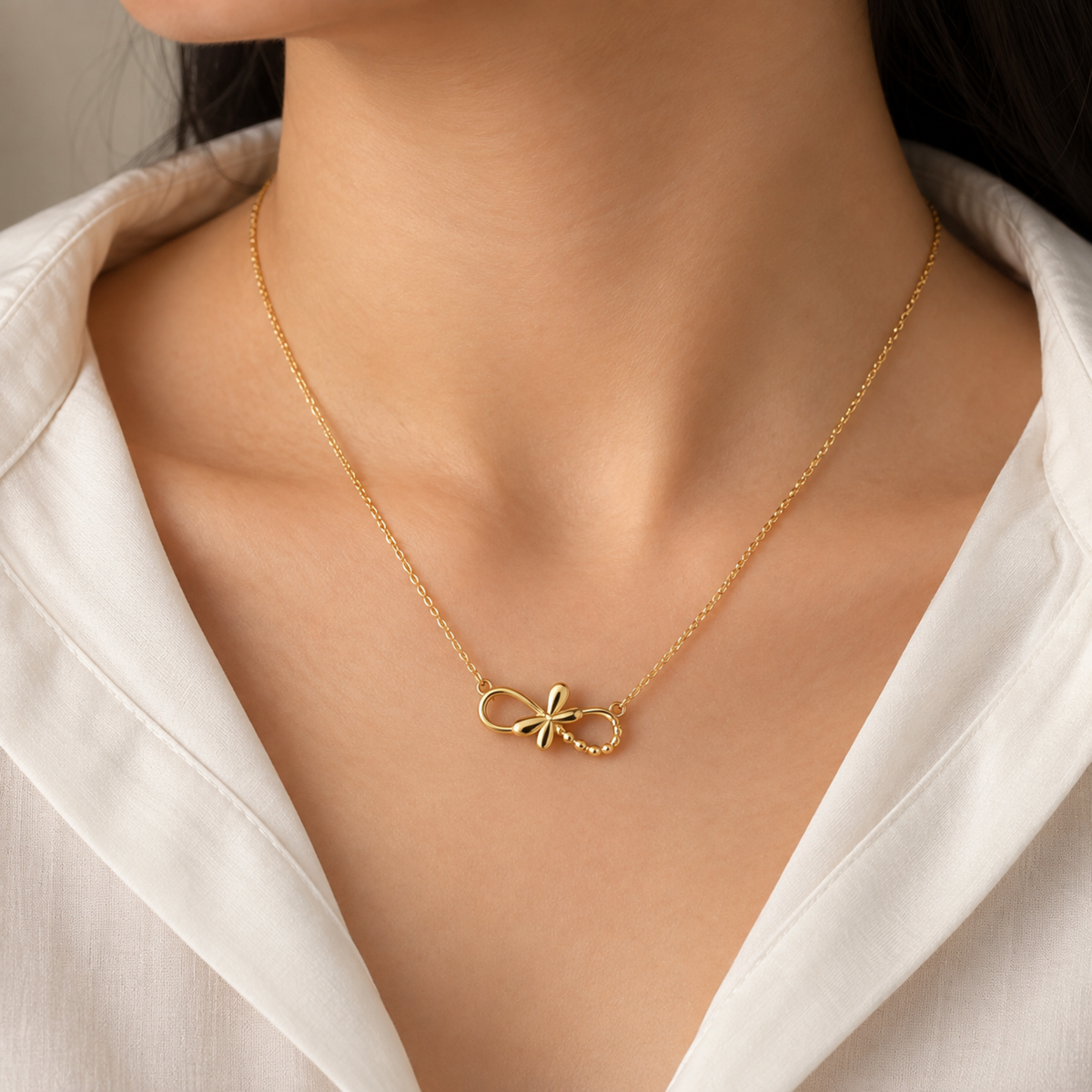 A close-up of a model wearing a delicate gold chain necklace with a small bow-shaped pendant, styled with a white collared shirt.