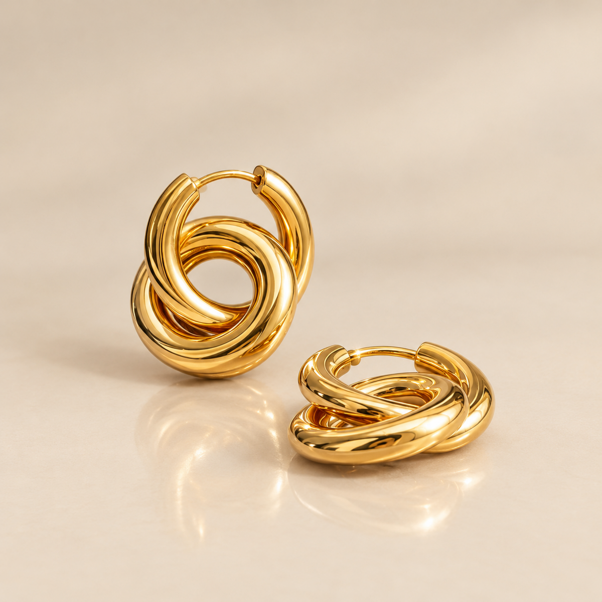 A pair of polished gold huggie hoop earrings with a twisted knot design, displayed on a warm beige reflective surface.