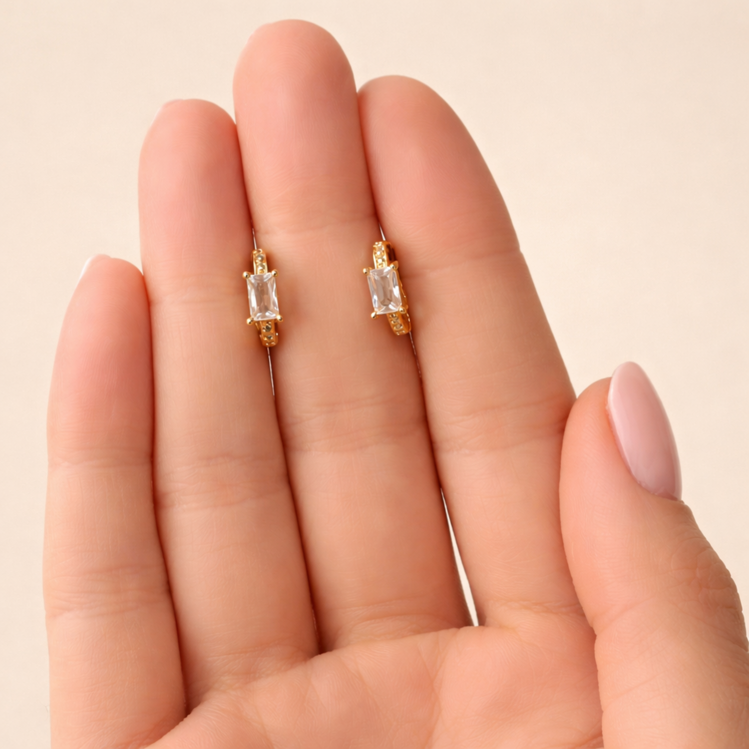 A pair of gold-plated huggie hoop earrings for women featuring a central baguette-cut clear crystal, shown on a hand