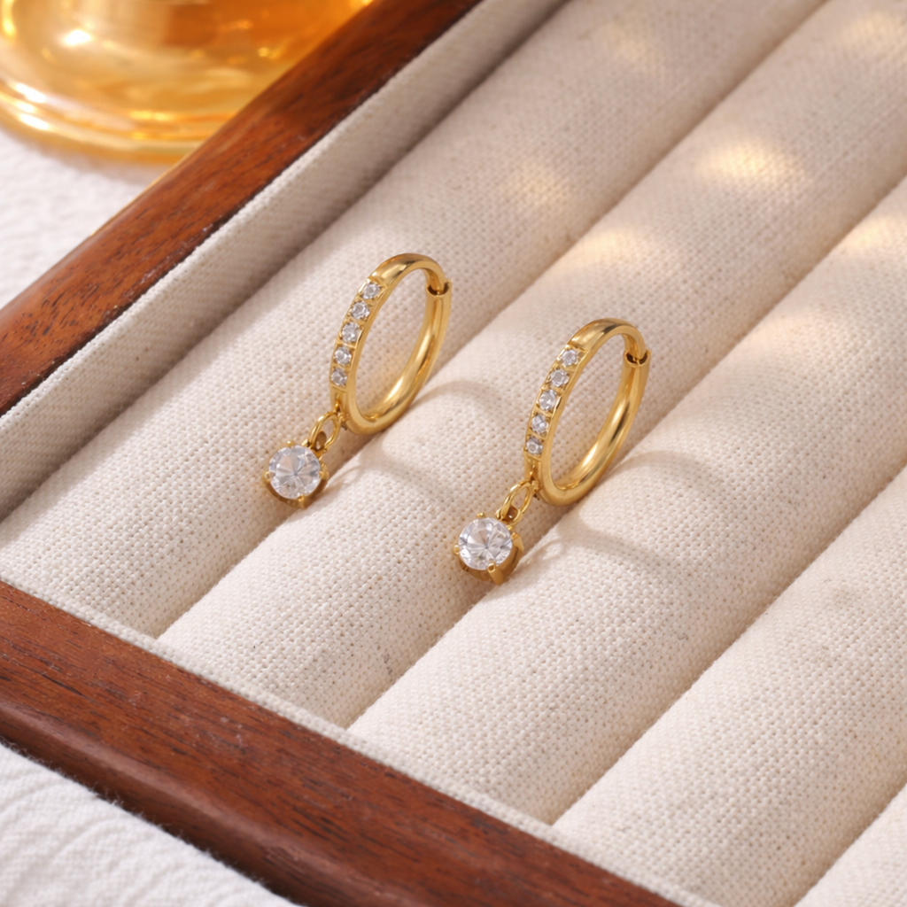 A pair of gold-plated hoop earrings for women featuring a sparkling round-cut crystal drop, shown on a jewellery organizer