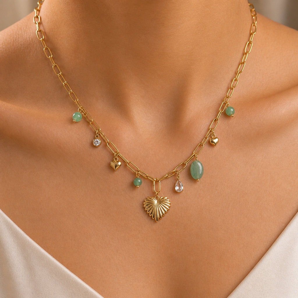 A gold-plated stainless steel chain for women featuring paperclip links and a variety of charms including a textured gold heart, green beads, and crystal accents, shown on a model