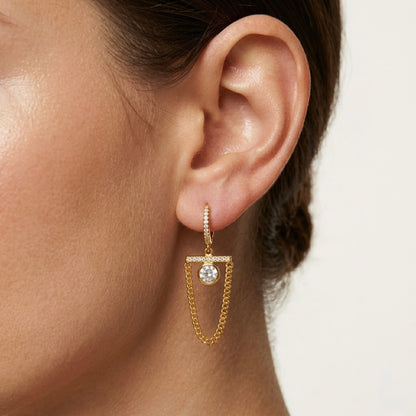 A pair of gold-plated long earrings for women featuring a hoop, a horizontal crystal bar, and a dangling gold curb chain loop, shown on a model