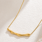 A close-view of a gold-plated stainless steel chain for women featuring a horizontal, fluid-shaped "liquid gold" ripple bar pendant on a sleek snake chain