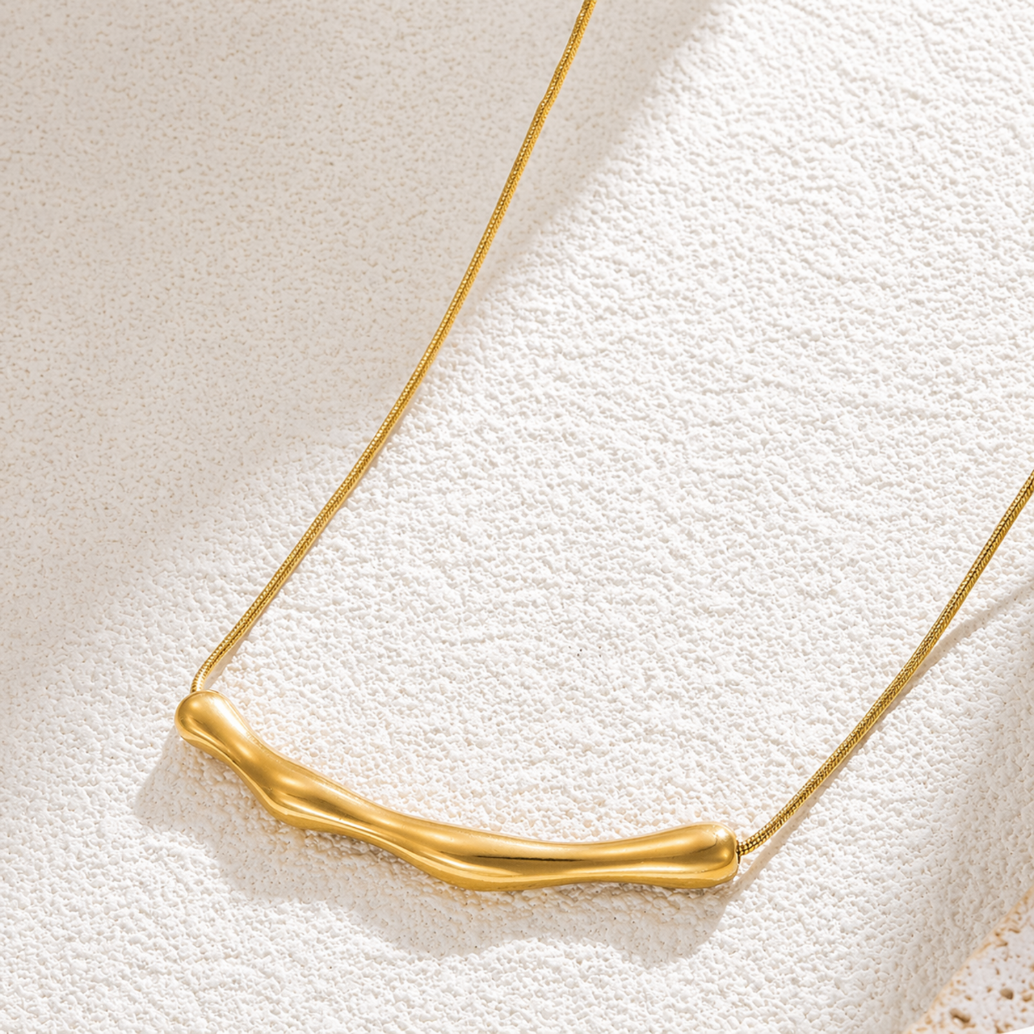 A close-view of a gold-plated stainless steel chain for women featuring a horizontal, fluid-shaped "liquid gold" ripple bar pendant on a sleek snake chain