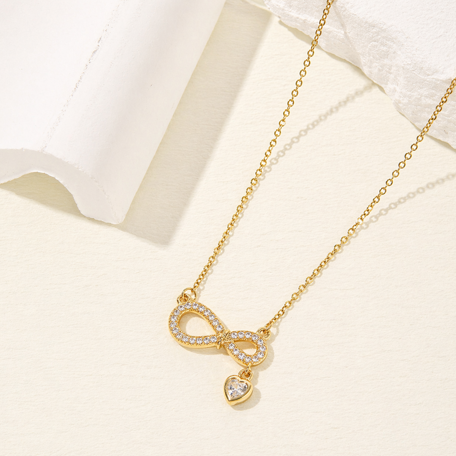 A gold-plated stainless steel chain for women featuring an infinity symbol pendant set with sparkling crystals and a small dangling heart-shaped crystal charm.