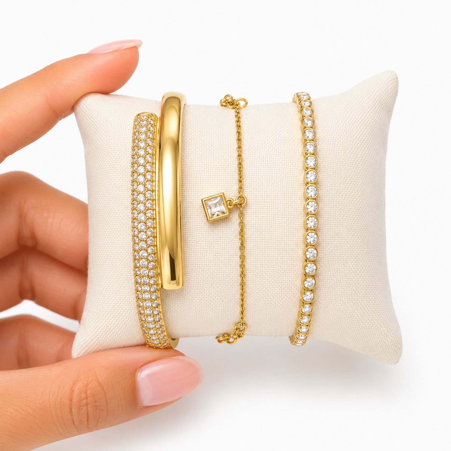 A close-view of a premium three-piece gold-plated bracelet stack for women featuring a thick half-pavé crossover bangle, a sleek bezel-set crystal bangle, and a fine chain bracelet with three dangling square crystal charms.