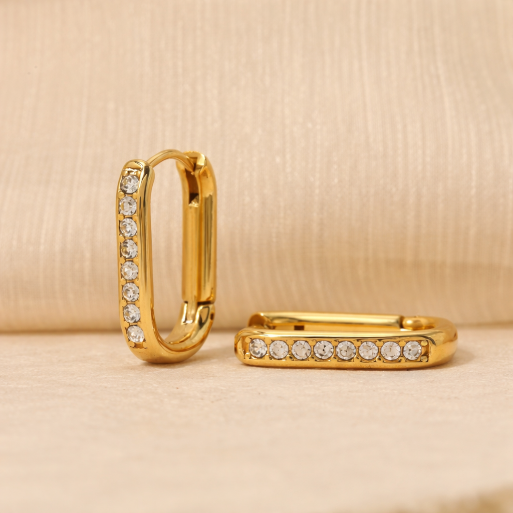 A pair of gold-plated rectangular hoop earrings for women featuring a vertical row of clear crystals, shown on a beige background