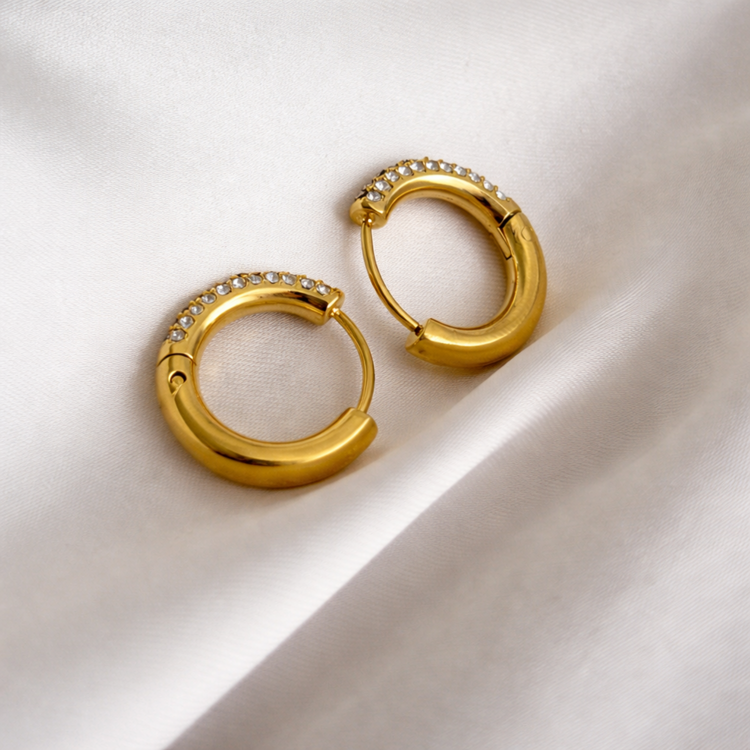 A pair of gold-plated thick hoop earrings for women featuring a front half encrusted with clear pavé crystals, shown on a cream cloth.