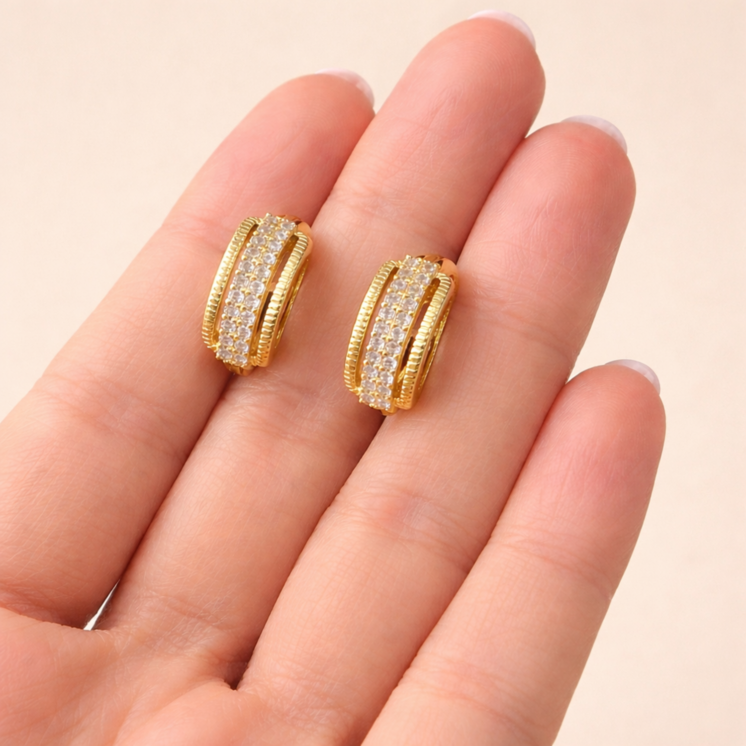 A pair of gold-plated textured hoop earrings for women featuring a central row of clear crystals and ridged outer bands, shown held in a hand