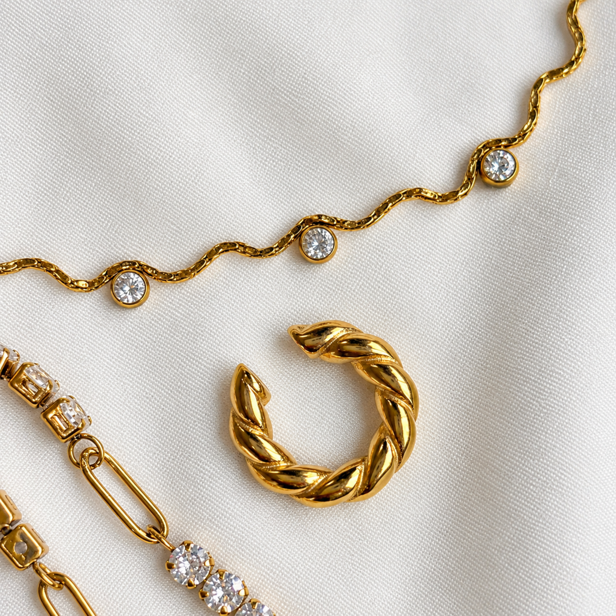 Gold jewelry set on a textured white fabric background, featuring a twisted rope ear cuff, a wavy chain necklace with round clear stones, and a gold paperclip chain with diamond accents.
