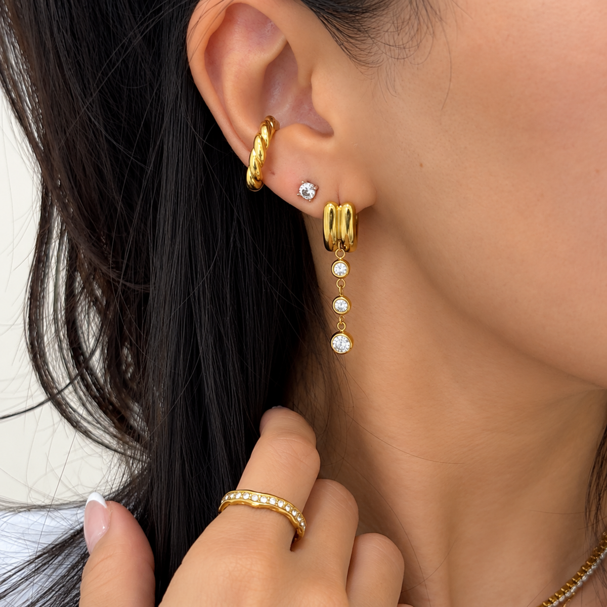 Close-up of a model wearing gold hoop earrings, a small stud earring, and a gold dangle earring with clear gemstone accents, along with a gold ear cuff.