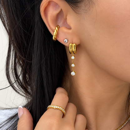 Close-up of a model wearing gold hoop earrings, a small stud earring, and a gold dangle earring with clear gemstone accents, along with a gold ear cuff.