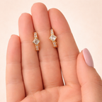 A pair of gold-plated huggie hoop earrings for women featuring a central kite-set princess-cut clear crystal, shown on a hand