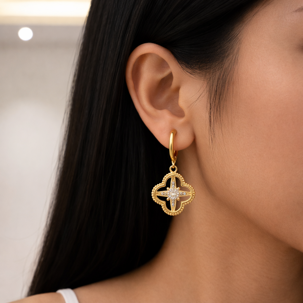 A gold-plated hoop earrings for women featuring a dangling clover-shaped lattice charm with a central crystal stone, shown on a model