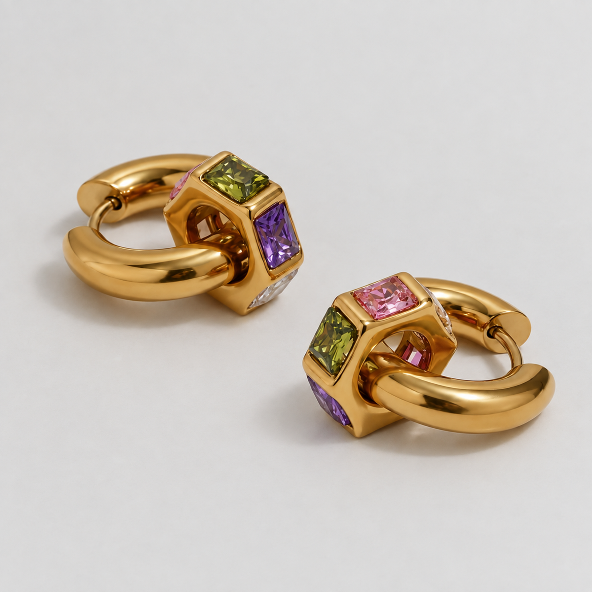 A pair of gold huggie hoop earrings with a central cube-shaped bezel set with colored gemstones, including green, purple, and pink, on a light gray background.