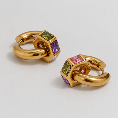 A pair of gold huggie hoop earrings with a central cube-shaped bezel set with colored gemstones, including green, purple, and pink, on a light gray background.