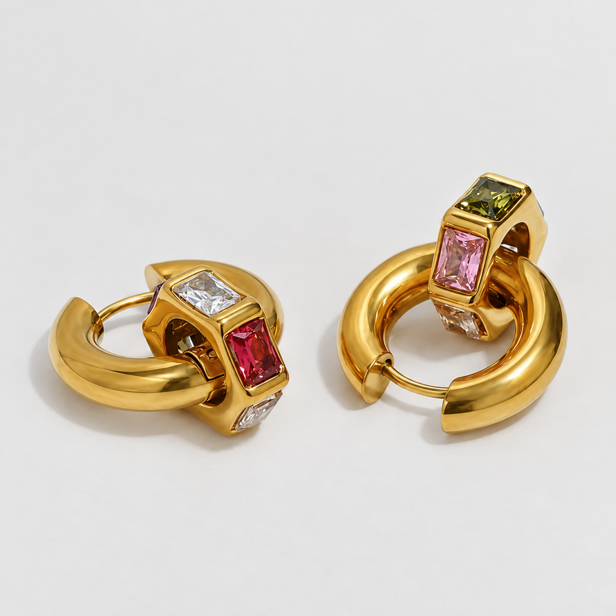 A pair of gold huggie hoop earrings with rectangular gemstone accents in pink, red, green, and clear, displayed on a light gray background.