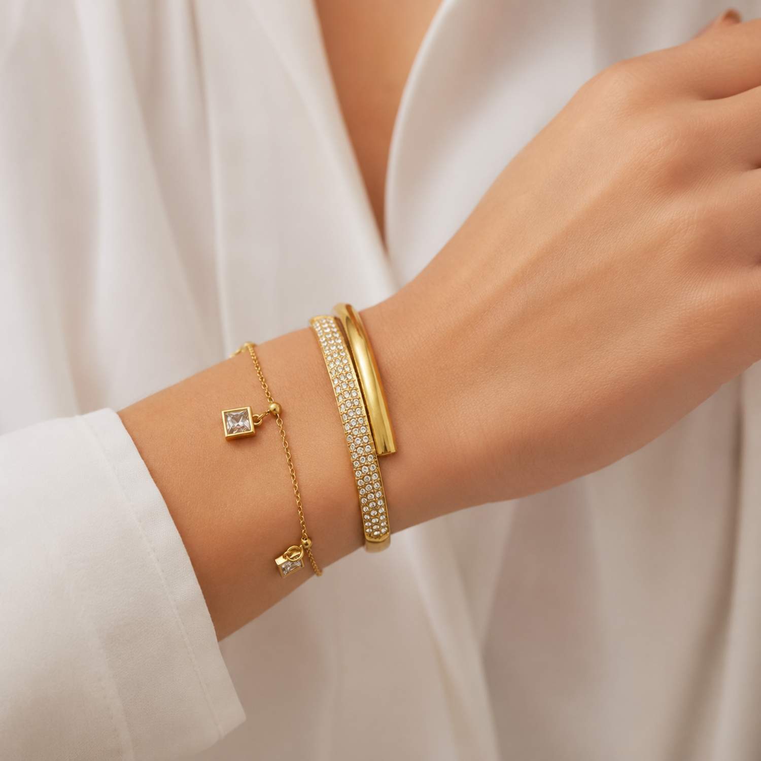 A premium two-piece gold-plated bracelet stack for women featuring a thick half-pavé crossover bangle and a fine chain bracelet with three dangling square crystal charms on a model