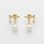 A close-view of a pair of gold-plated arc drop earrings for women featuring a central white pearl and a small clear crystal, shown on a white background
