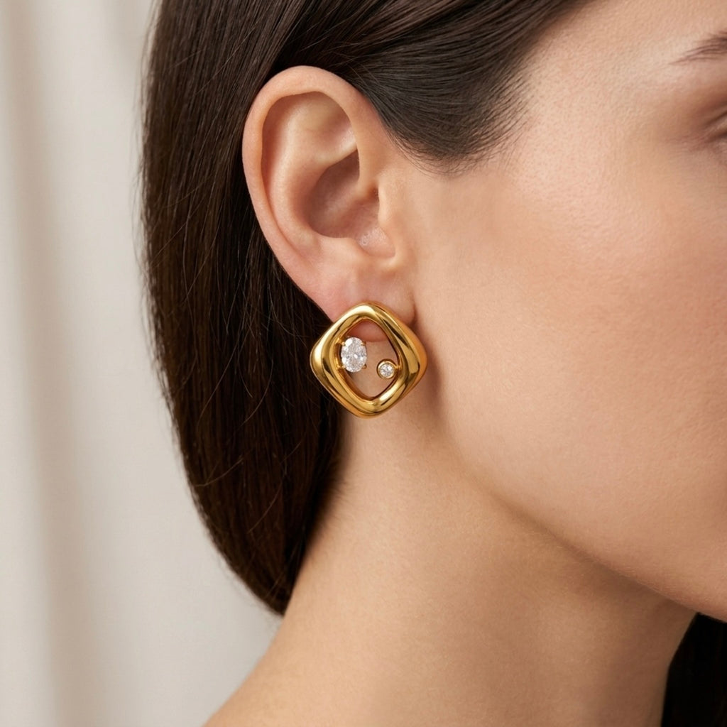 A gold-plated abstract rhombus-shaped stud earrings for women featuring two floating crystals inside a molten gold frame, shown on a model