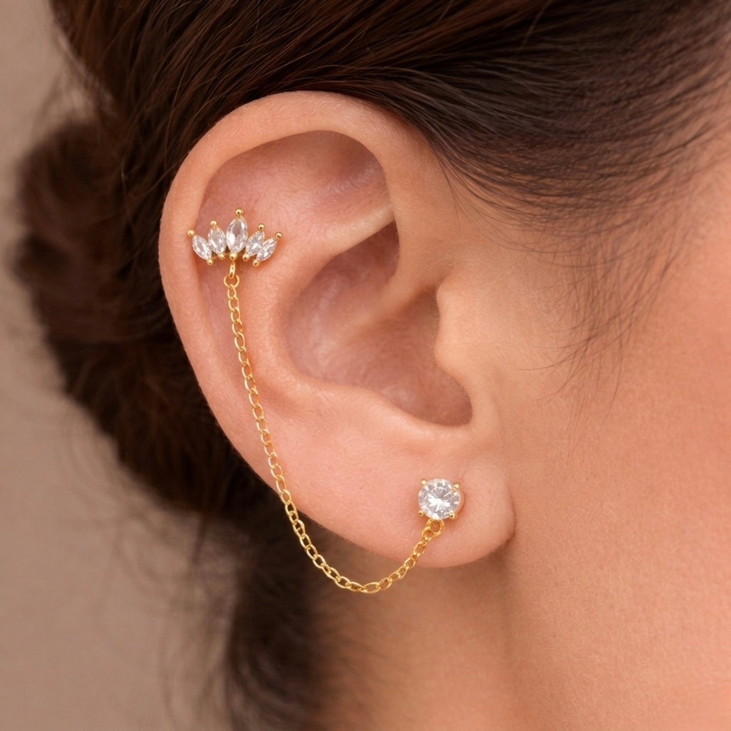 A gold-plated ear chain earrings for women featuring a five-stone marquise crown stud and a round white stone stud connected by a delicate gold chain on a model
