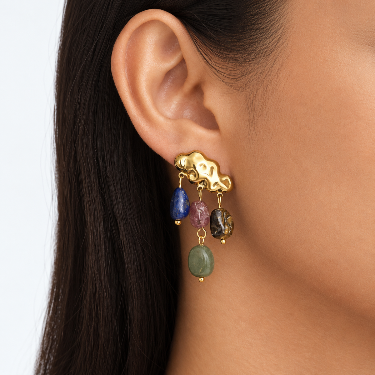 A close-up of a model's ear wearing a gold ear cuff with dangling multicolor gemstone charms.