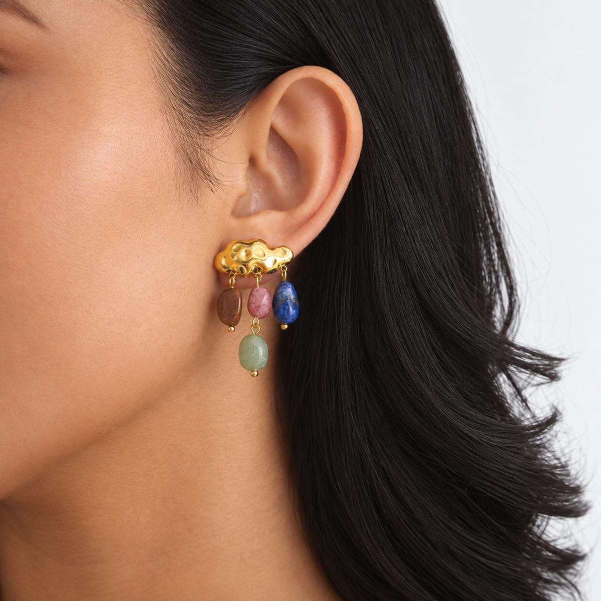 A close-up of a model's ear wearing a gold cloud-shaped dangle earring with five colorful dangling beads in brown, pink, blue, green, and gold.