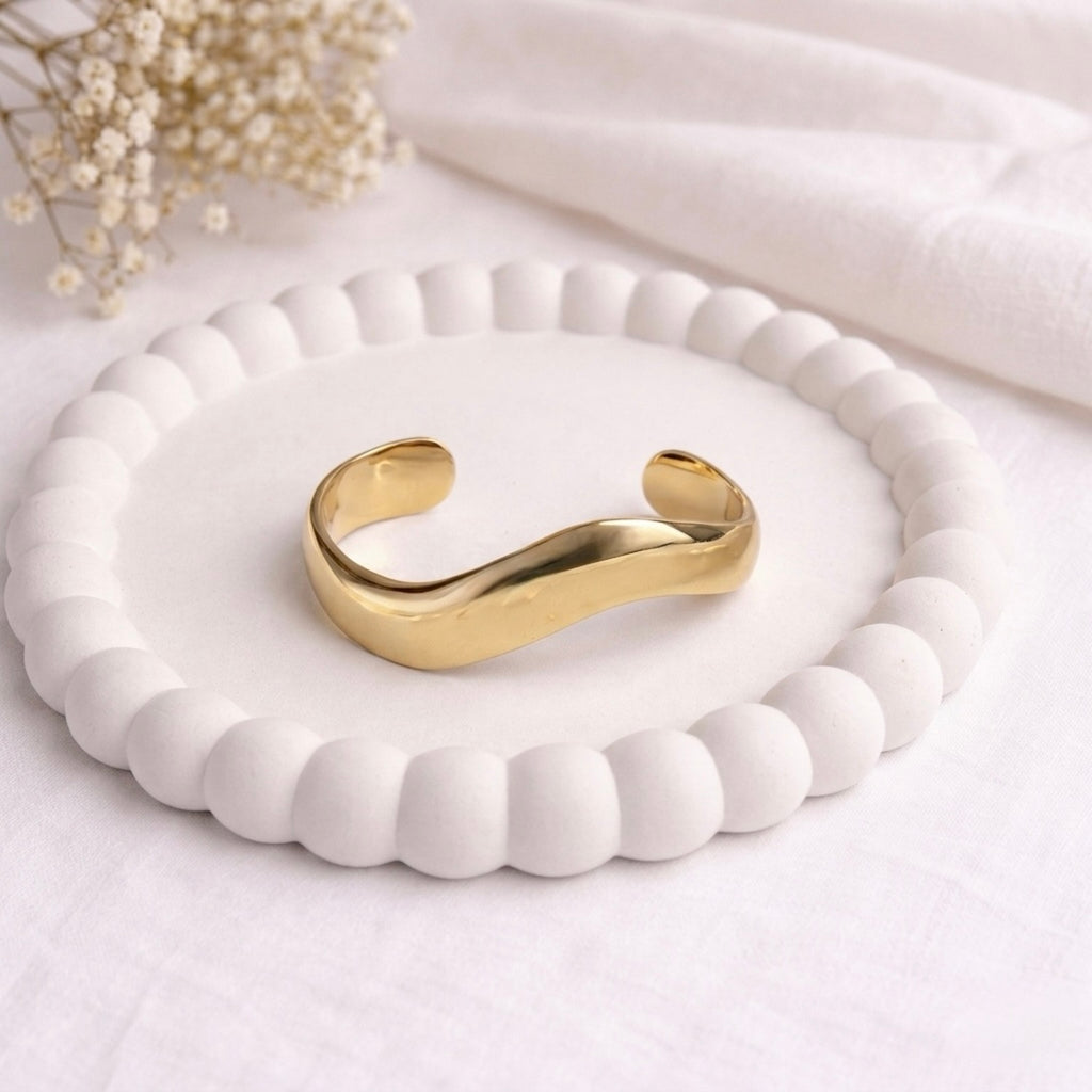 A bold and wide gold-plated organic wave cuff bracelet for women with a smooth, thick finish, displayed on a white bobbled ceramic tray