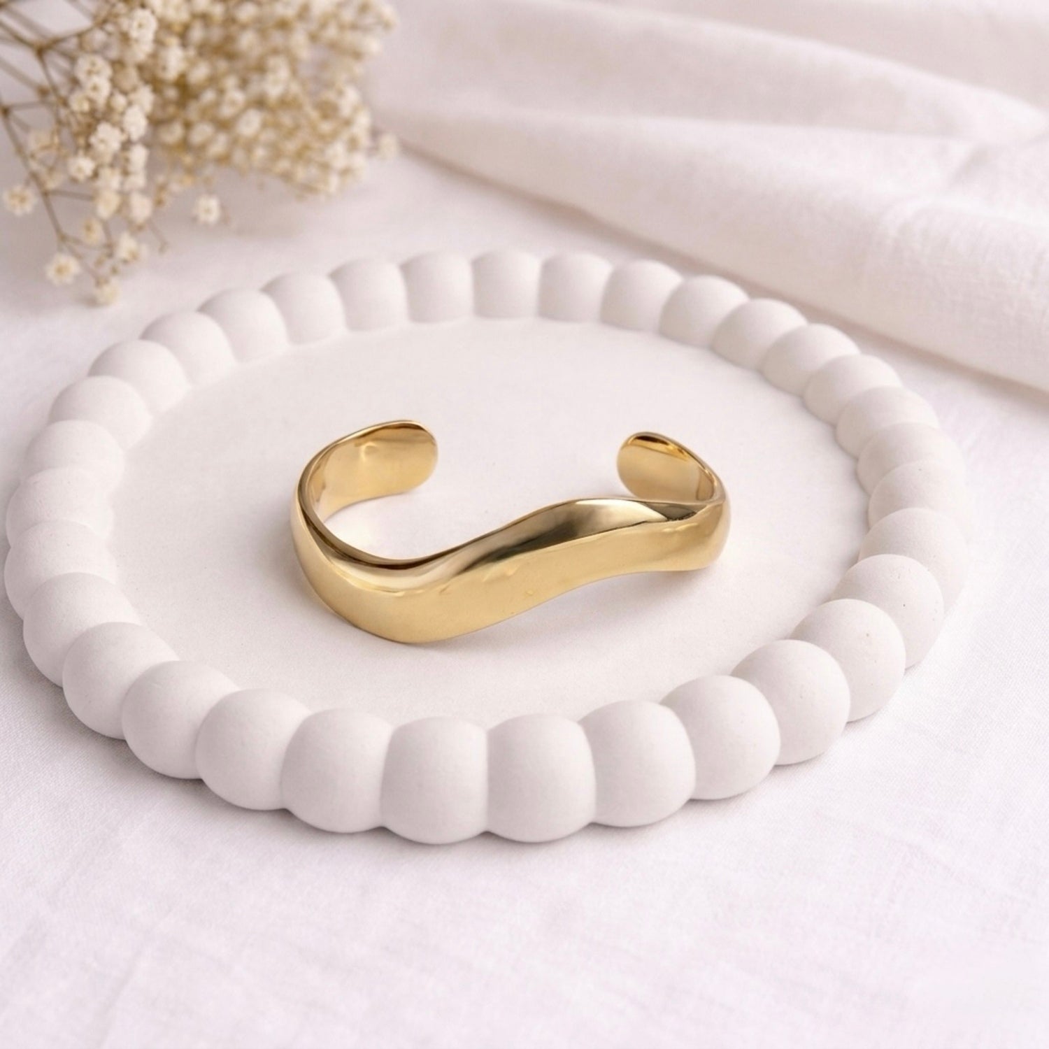 A bold and wide gold-plated organic wave cuff bracelet for women with a smooth, thick finish, displayed on a white bobbled ceramic tray