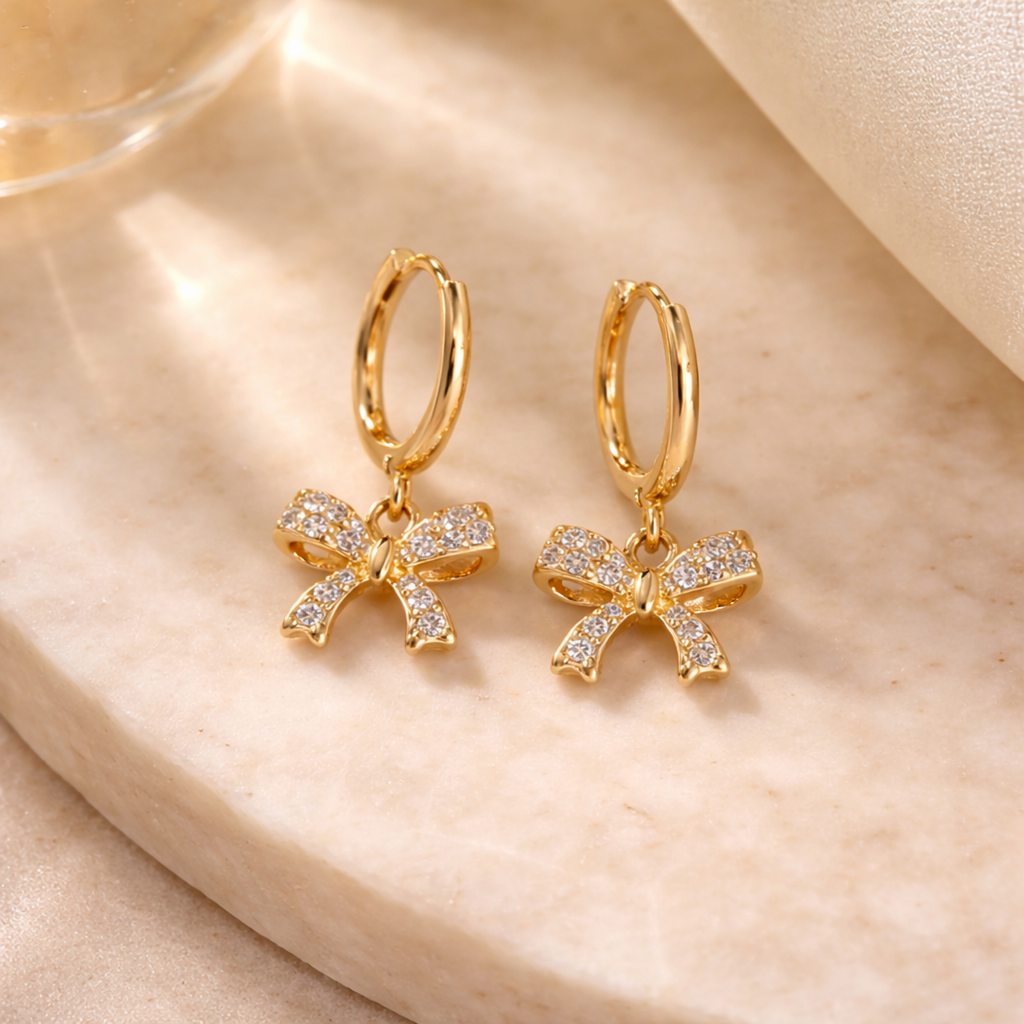 A gold-plated hoop earrings for women featuring a sparkling crystal-encrusted bow drop, shown on a beige background