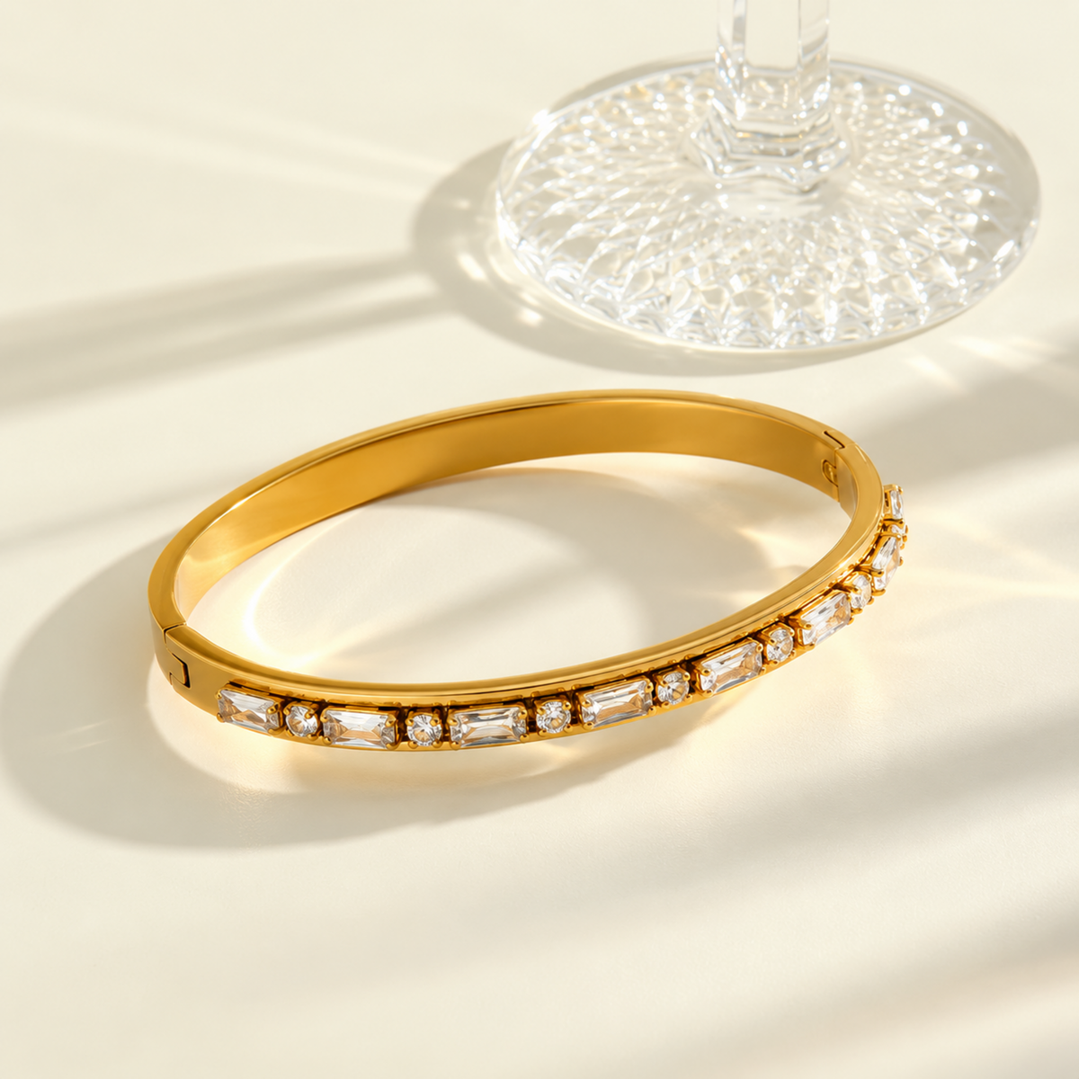 A gold bangle bracelet with a row of clear rectangular baguette and round crystals along the front, displayed on a warm cream surface with a glass base in the background.