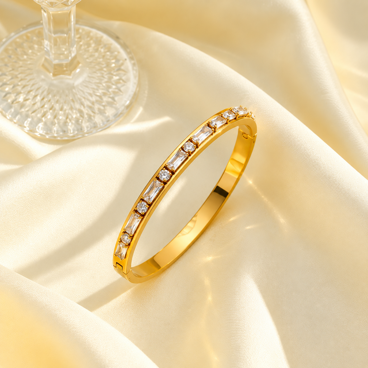 A gold bangle bracelet with a row of clear baguette and round stones, displayed on draped cream satin fabric next to a glass base.
